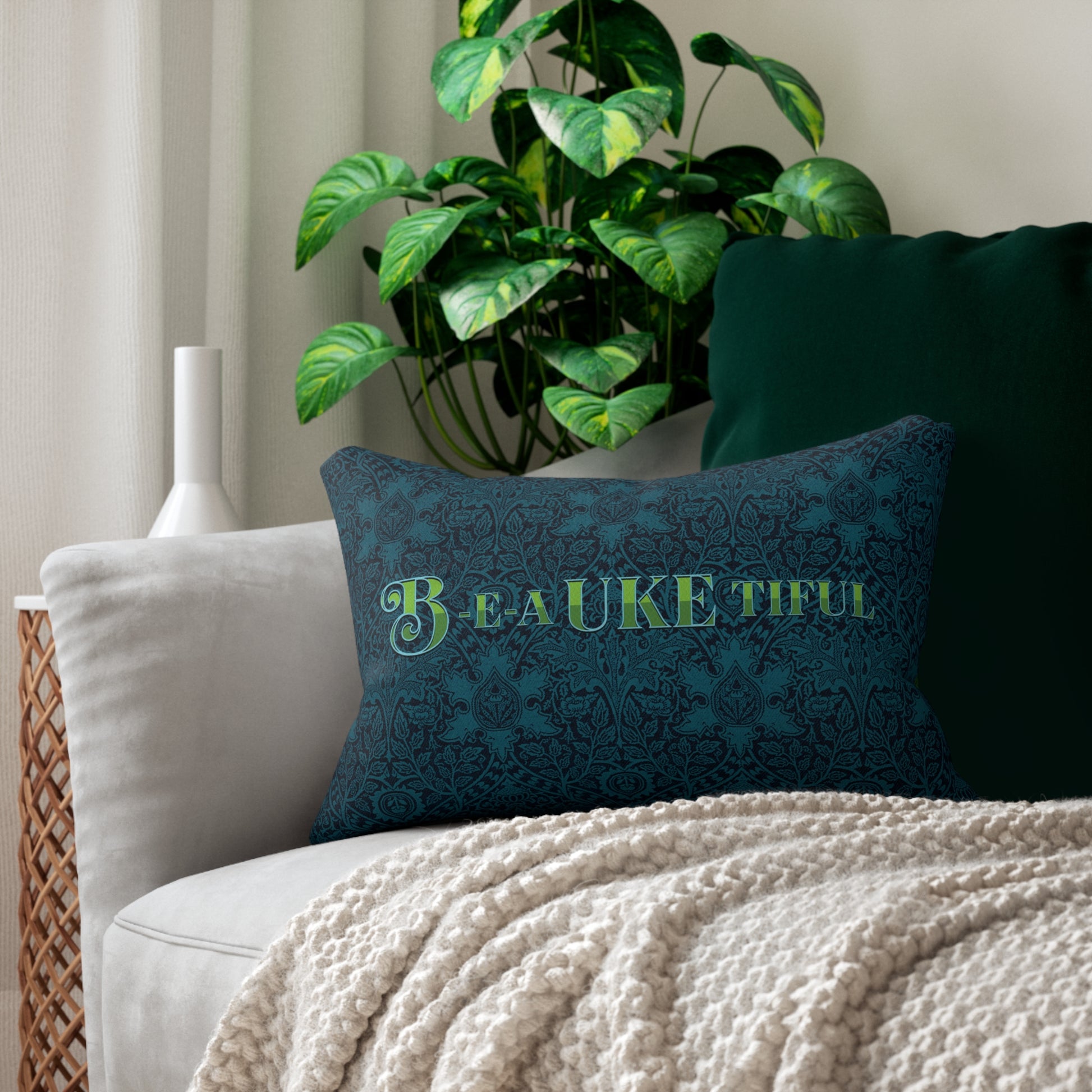 A throw pillow with a dark teal patterned background and 'B-E-A-UKE-tiful' in bold green letters printed on it, sitting on a sofa.