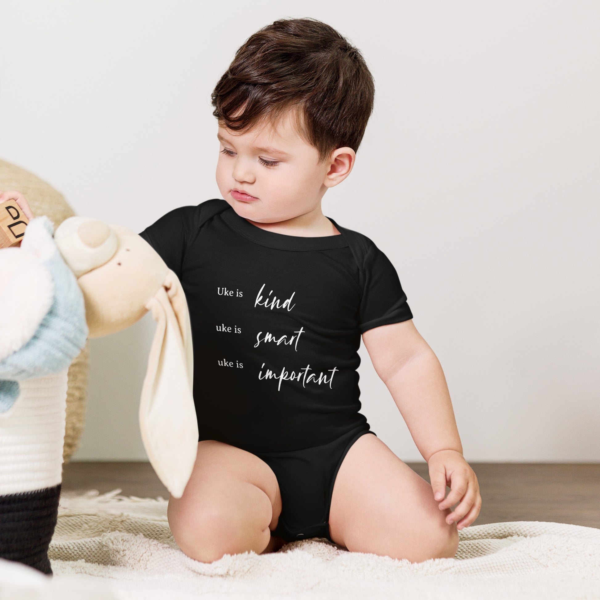 A baby wearing a black onesie printed with 'Uke is kind, uke is smart, uke is important' in white letters.