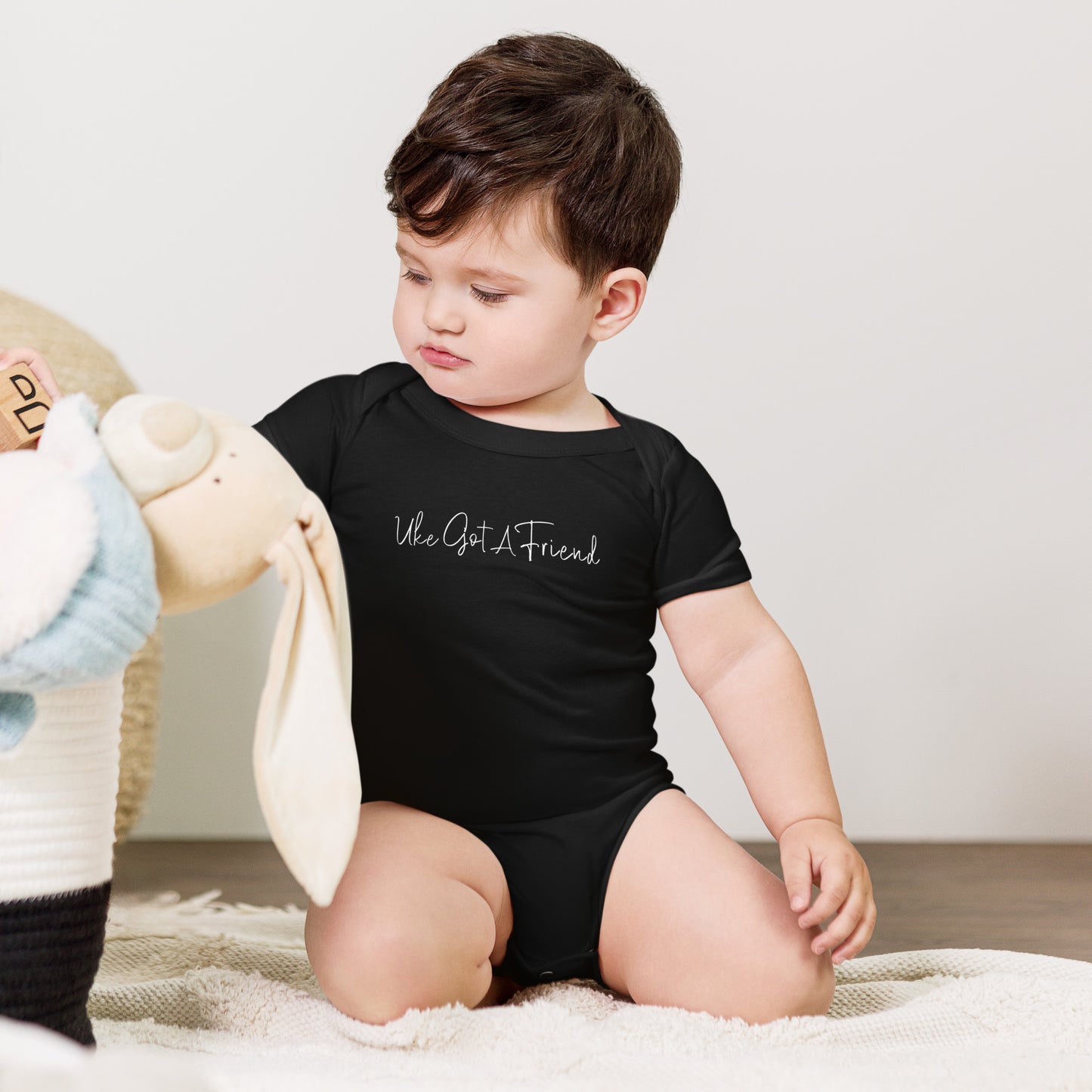 A baby wearing a black onesie printed with 'Uke Got A Friend' in white cursive text.