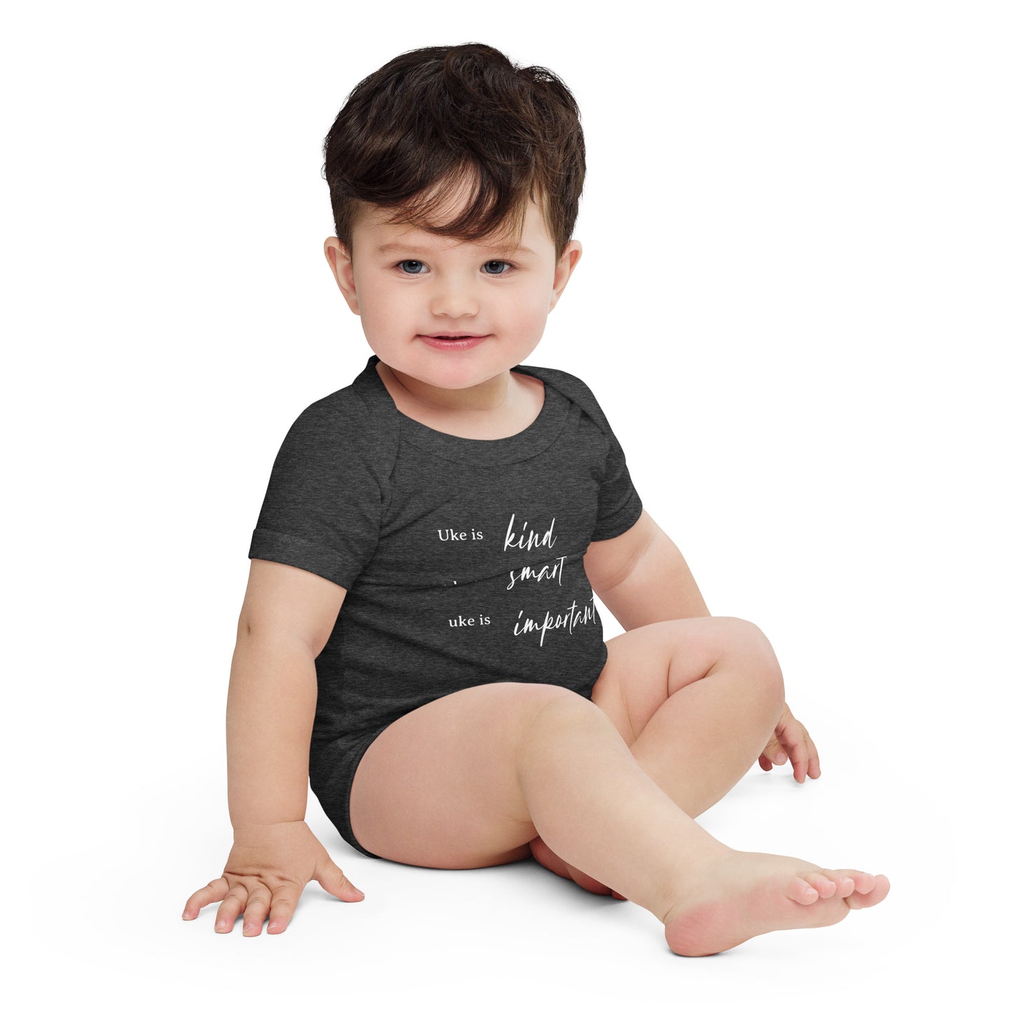 A baby wearing a dark grey onesie printed with 'Uke is kind, uke is smart, uke is important' in white letters.