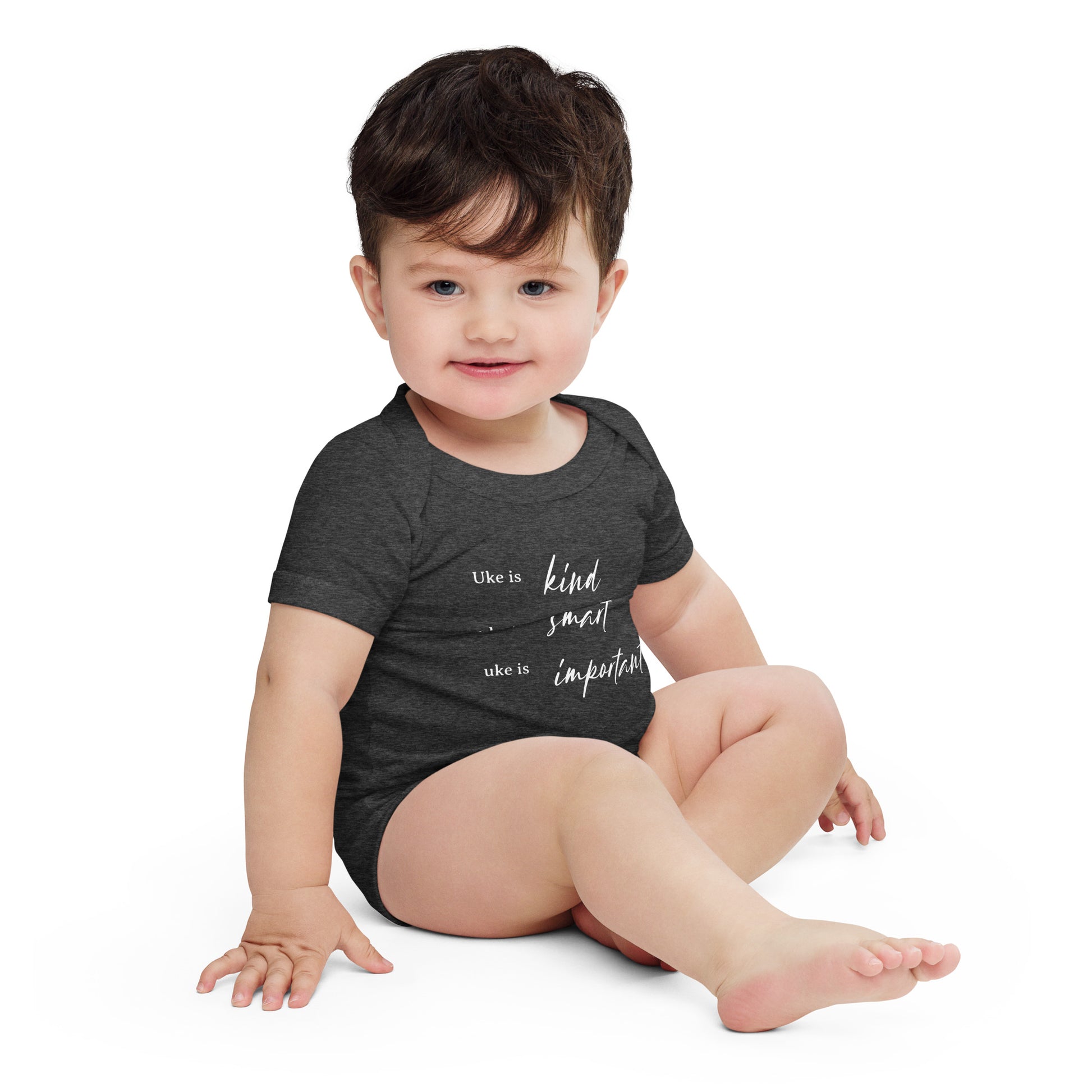 A baby wearing a dark grey onesie printed with 'Uke is kind, uke is smart, uke is important' in white letters.