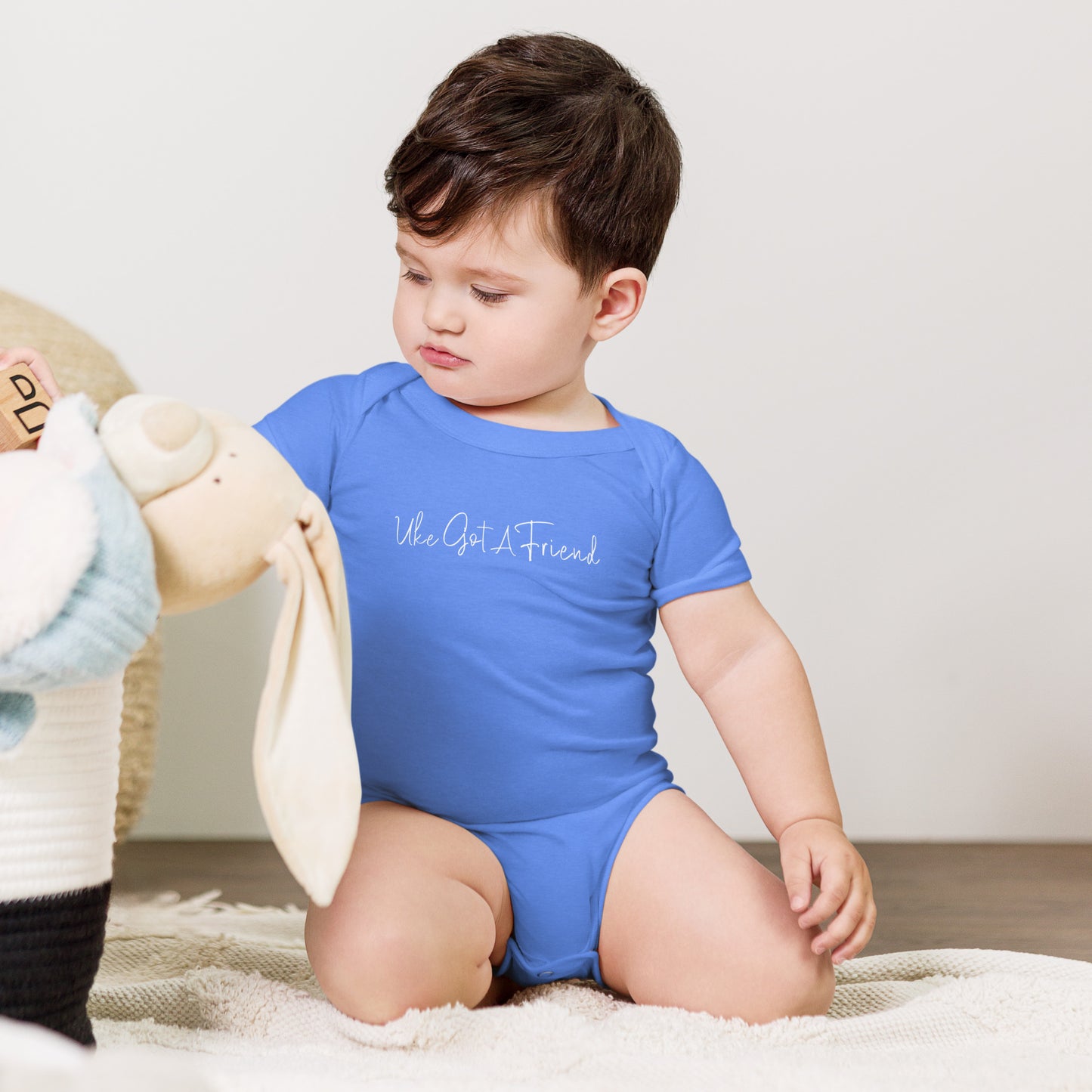 A baby wearing a heather Columbian Blue onesie printed with 'Uke Got A Friend' in white cursive text.