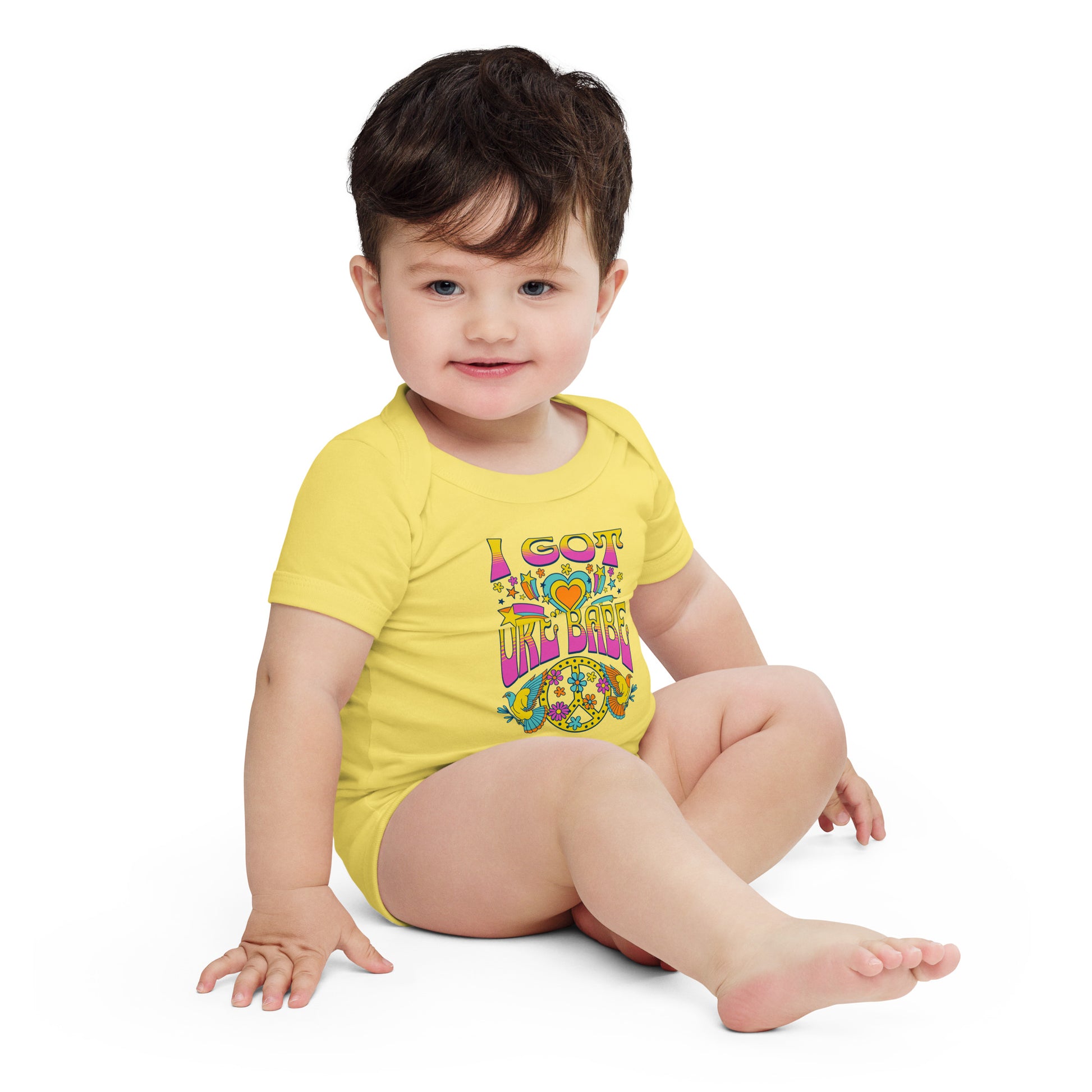 Baby wearing a yellow onesie printed with a pink, blue, and yellow 'I Got Uke Babe’ and peace sign graphic.