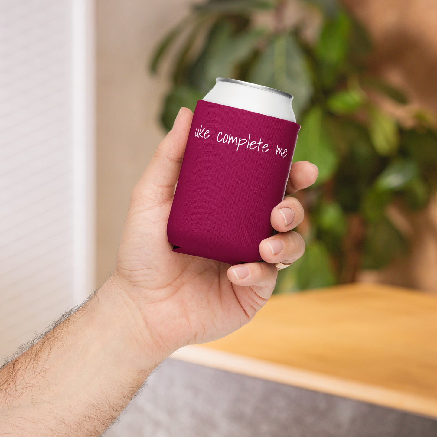 A hand holding a regular-sized can coozie printed with a maroon background and the words 'Uke Complete Me' in white lowercase text.
