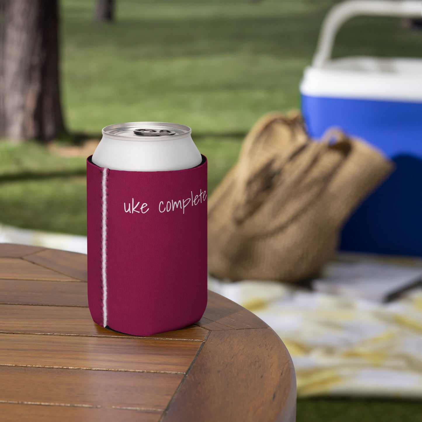 A hand holding a regular-sized can coozie printed with a maroon background and the words 'Uke Complete Me' in white lowercase text.