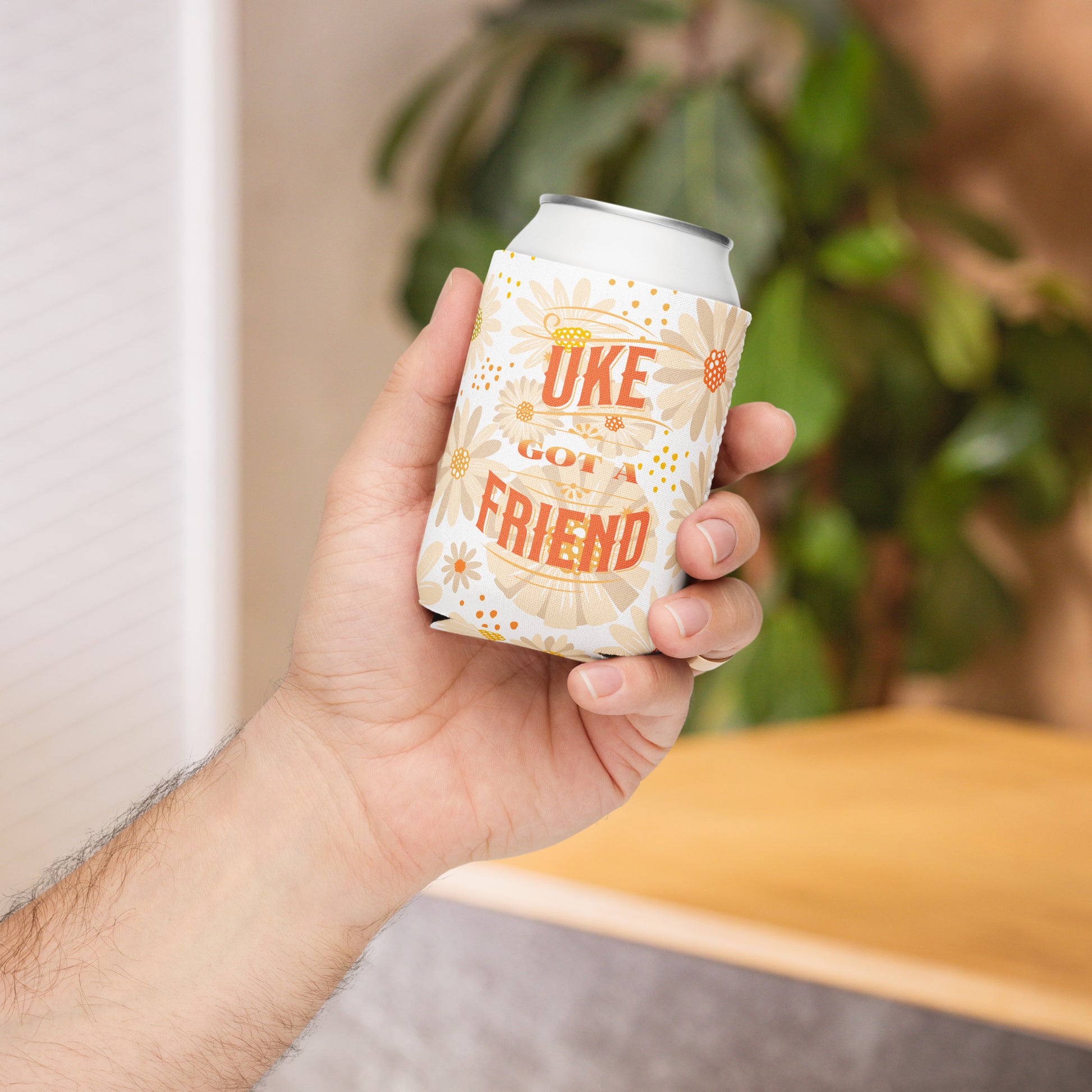 A hand holding a regular-sized can coozie with a flower design and the words 'Uke Got A Friend' in orange and yellow block letters printed on it.