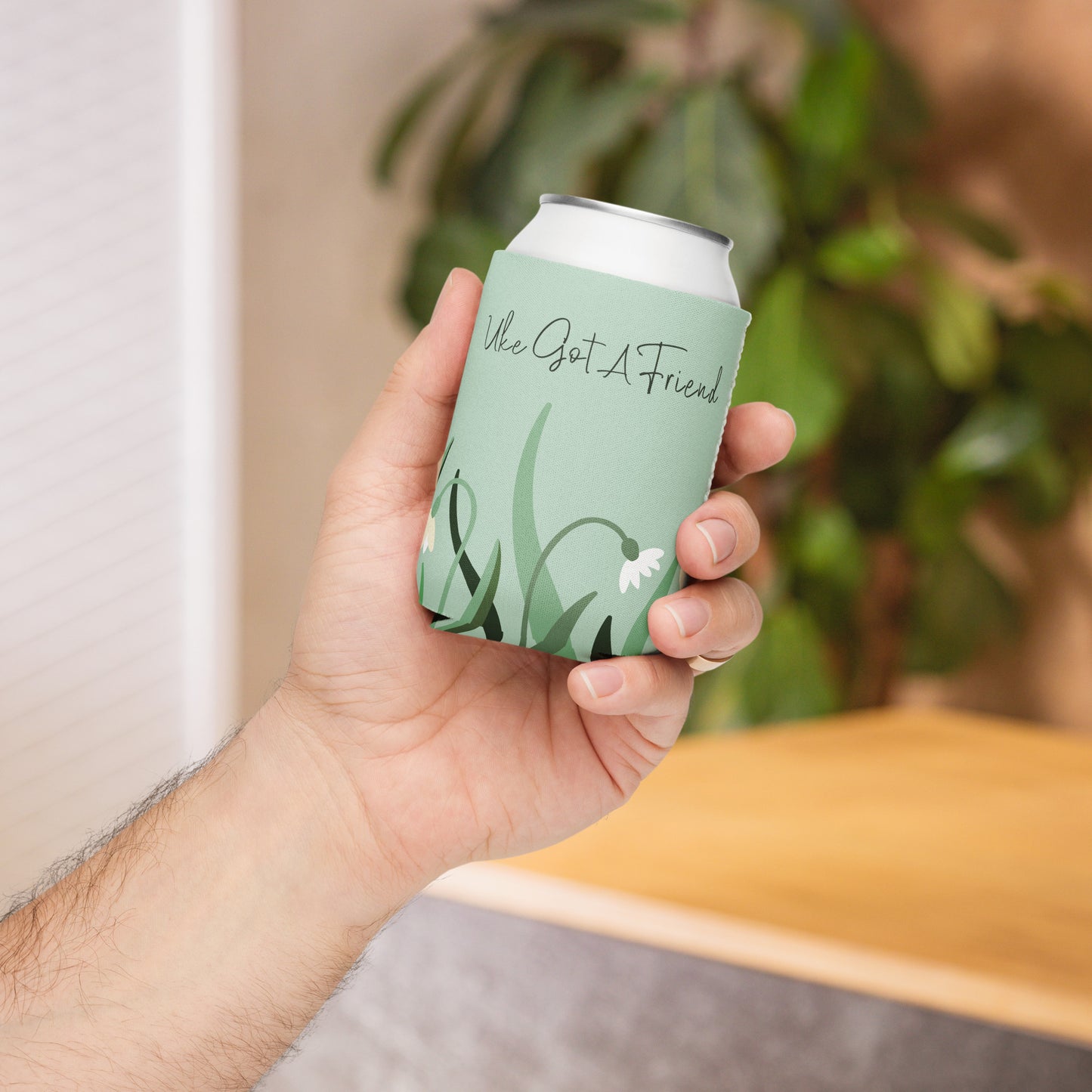 A hand holding a regular-sized can coozie with a flower design on a green background and the words 'Uke Got A Friend' printed on it.