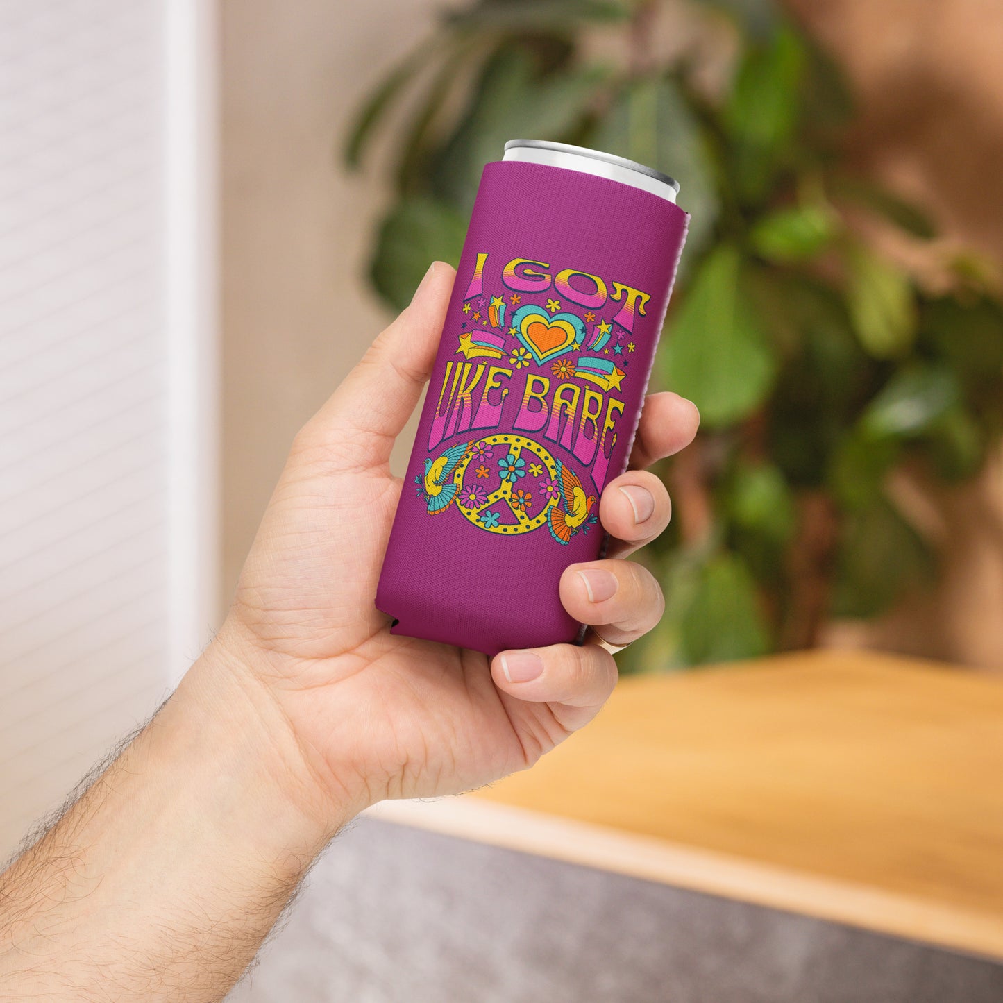 Hand holding a slim can coozie with a deep pink background and a pink, blue, and yellow 'I Got Uke Babe’ and peace sign graphic printed on it.