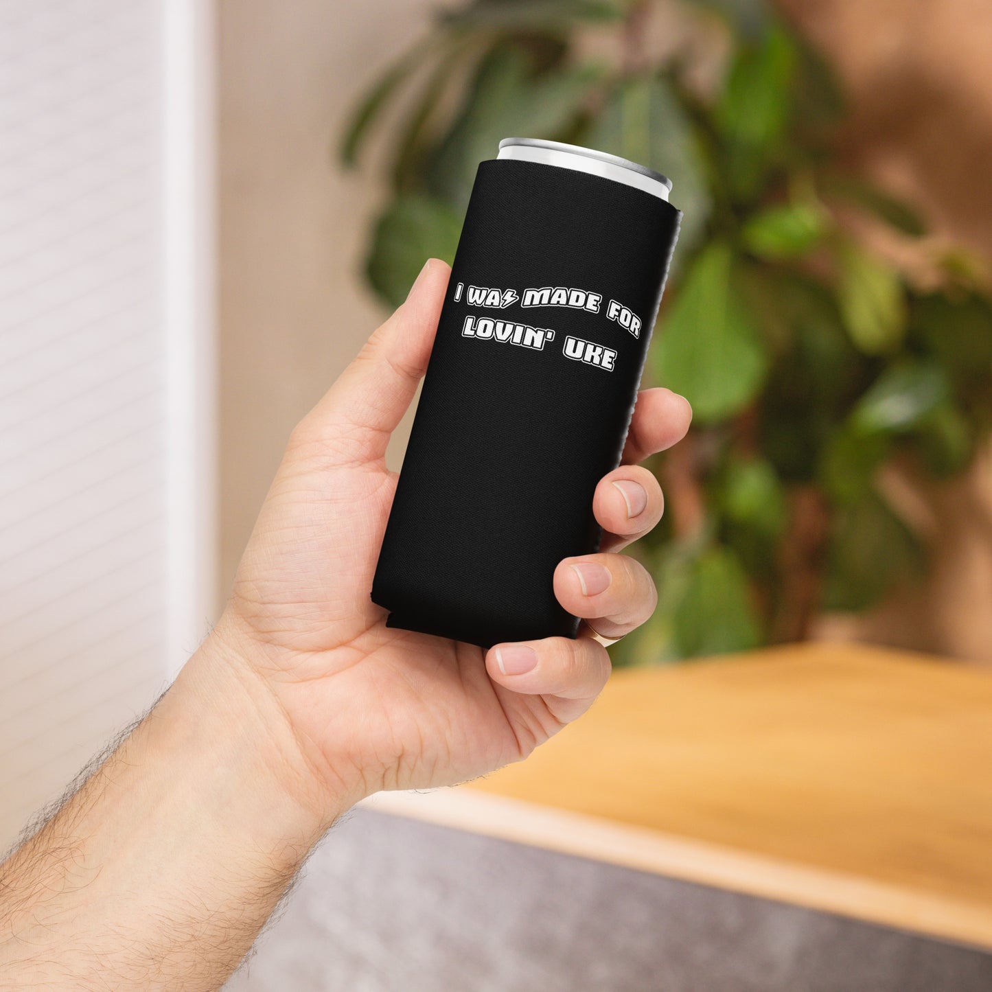 Hand holding a slim can coozie with a black background and 'I was made for lovin' uke' printed in black and white block letters.