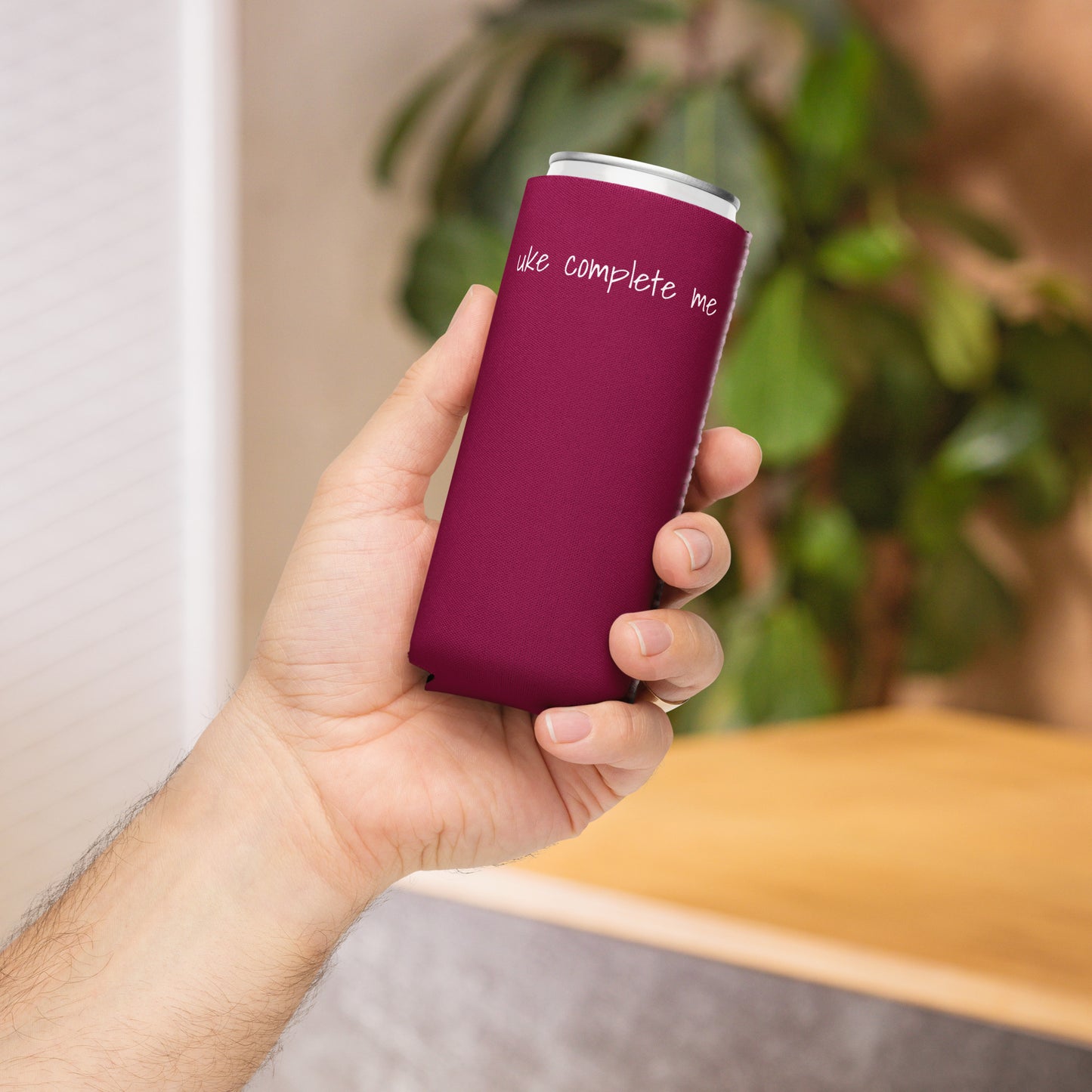 A hand holding a slim can coozie printed with a maroon background and the words 'Uke Complete Me' in white lowercase text.