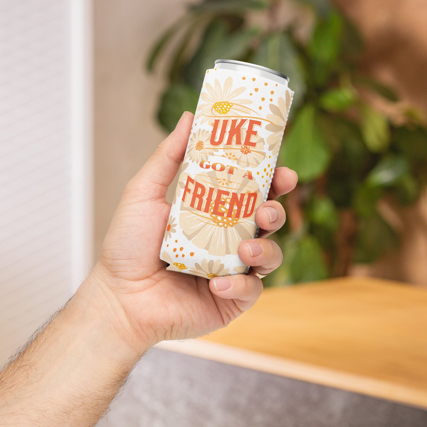 A hand holding a slim can coozie with a flower design and the words 'Uke Got A Friend' in orange and yellow block letters printed on it.
