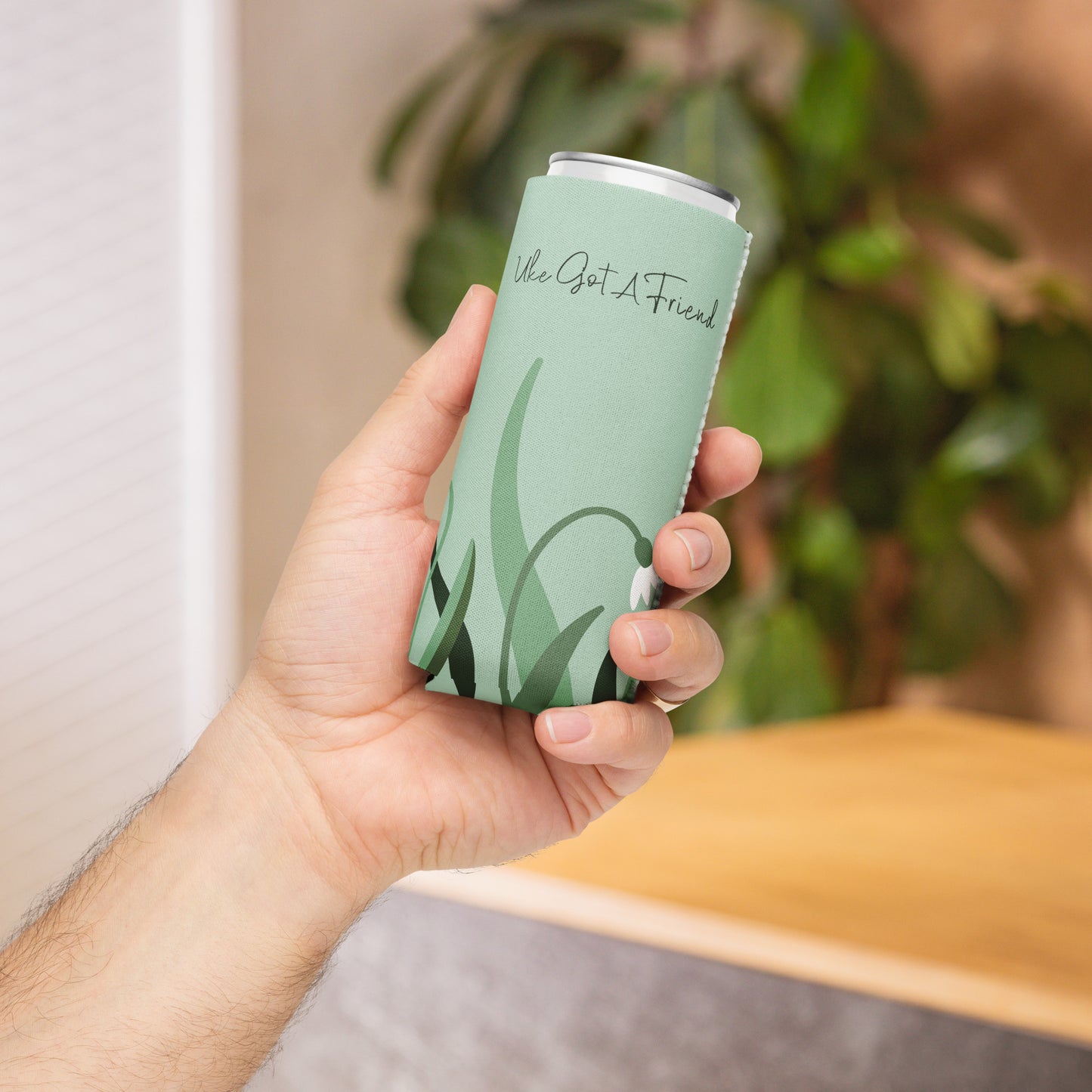 A hand holding a slim can coozie with a flower design on a green background and the words 'Uke Got A Friend' printed on it.