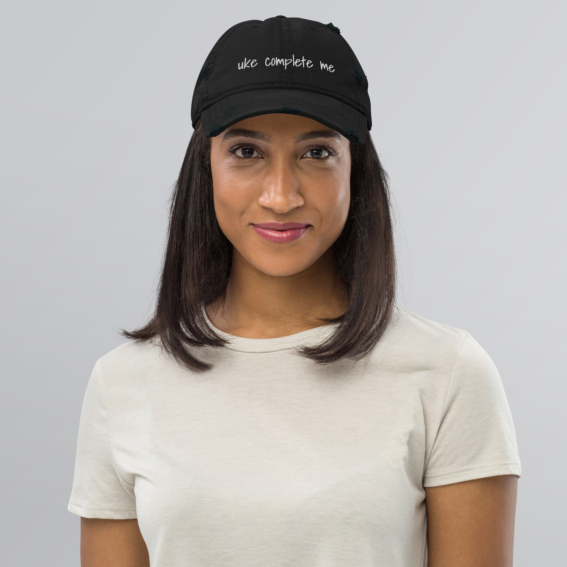 Woman wearing a black baseball cap with a distressed look and 'Uke complete me' embroidered on it with white thread.