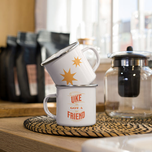 Two stacked enamel mugs showing the front and back of a single mug, with the words 'Uke Got A Friend' in orange and yellow block letters shown on one and two golden-colored stars on the other.