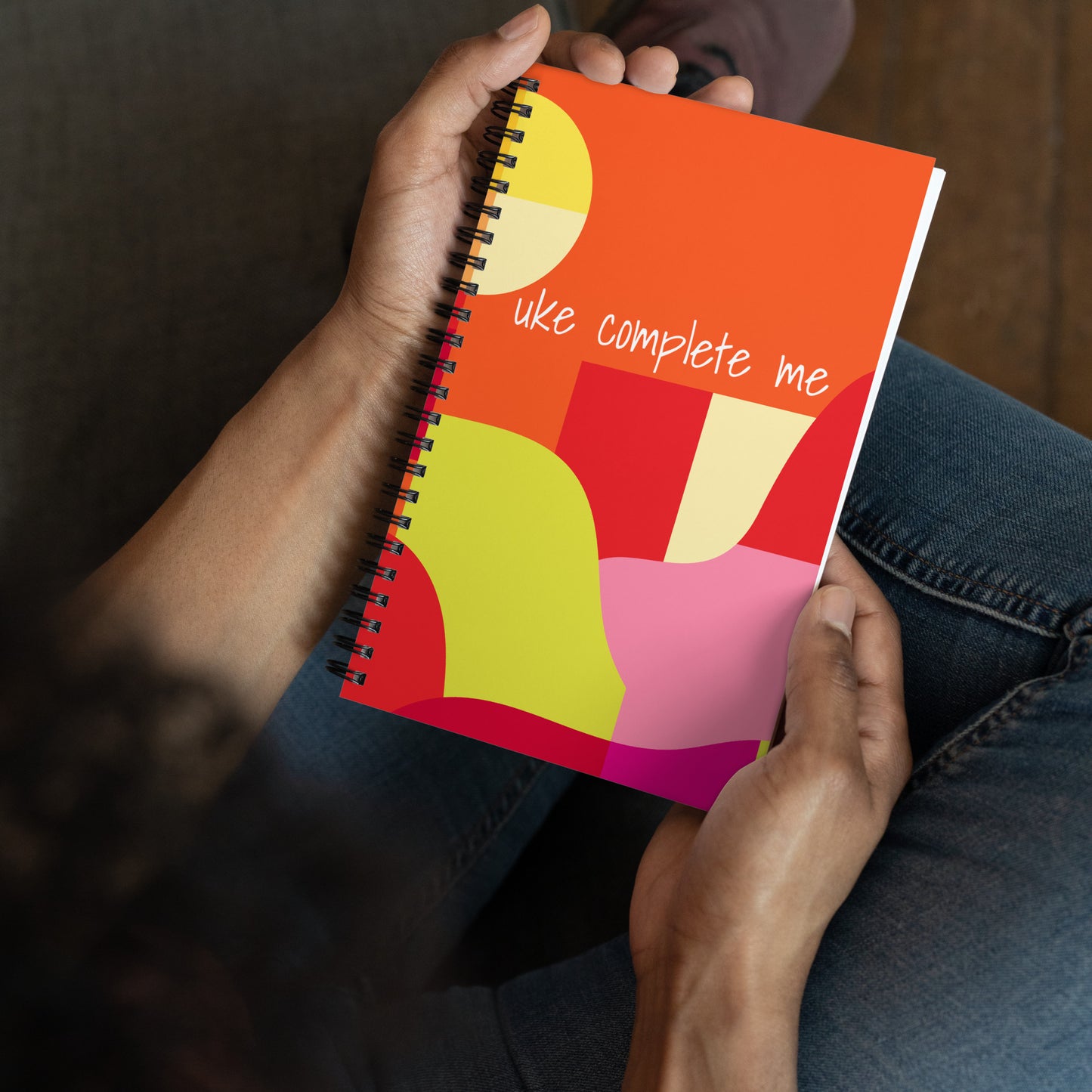 Hands holding a 5-by-7 inch notebook with 'uke complete me' in white text printed on a pink, yellow, and orange background.
