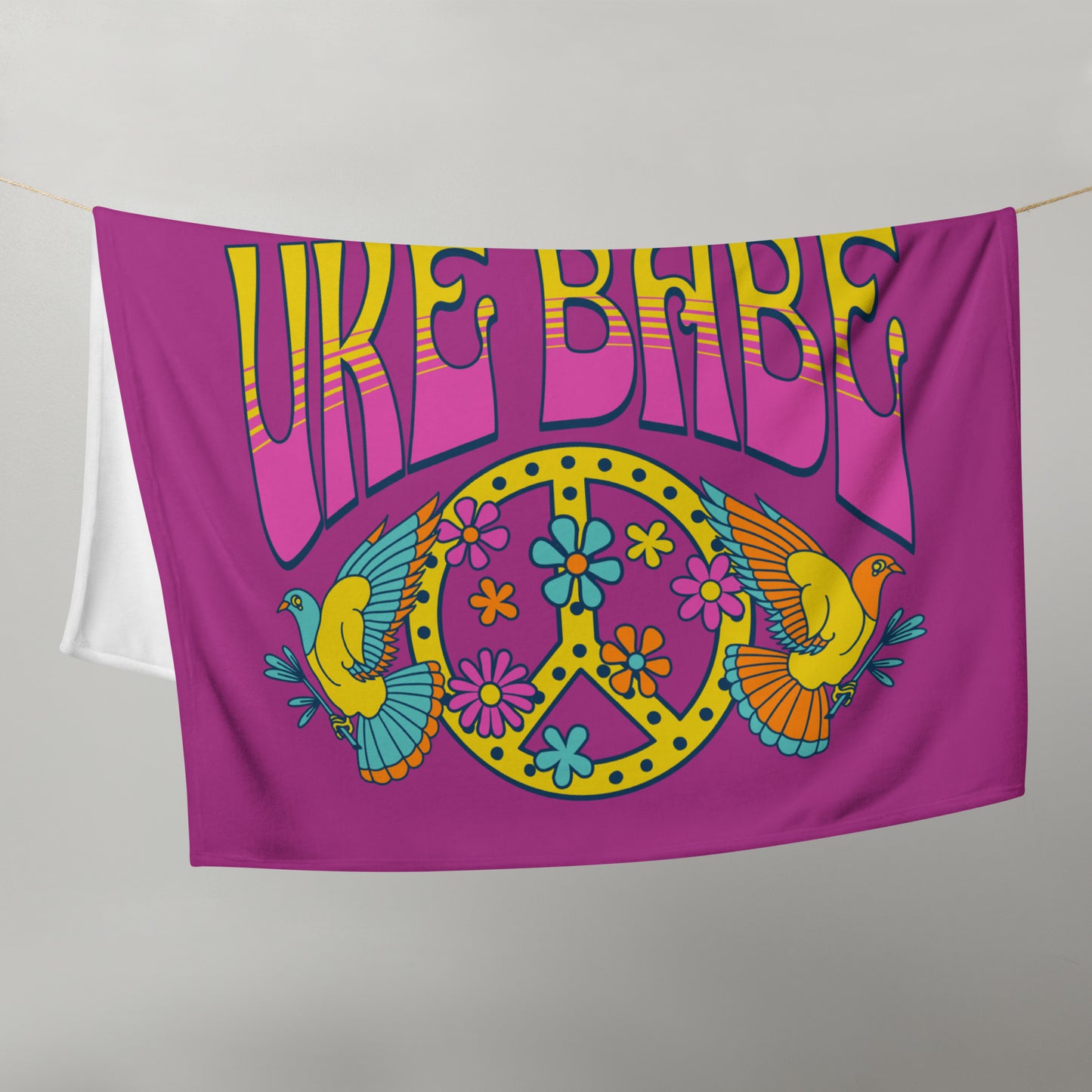 Microplush throw blanket with a dark pink background and a pink, blue, and yellow 'I Got Uke Babe' and peace sign graphic printed on it, draped over a clothesline.