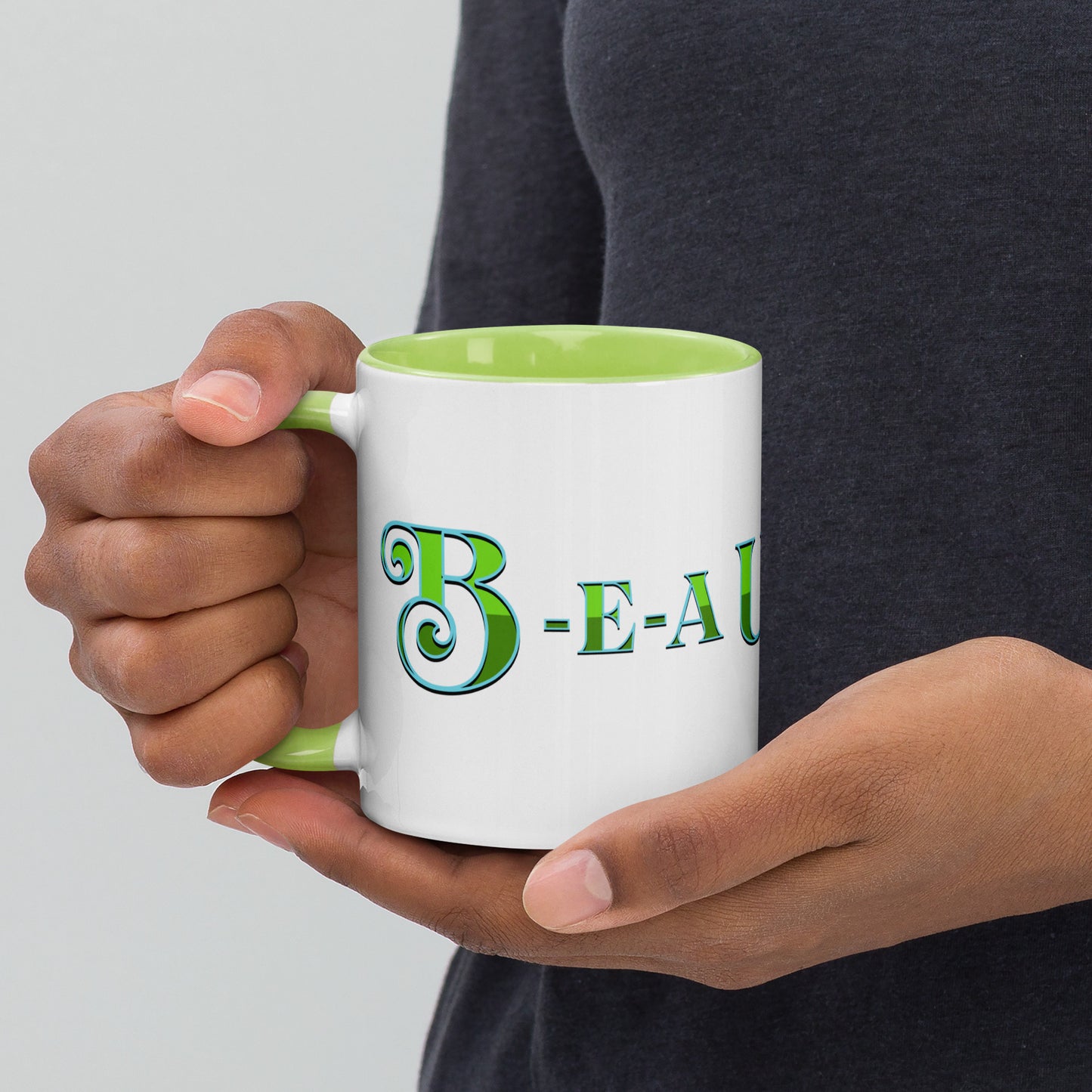 A person holding a white mug with light green handle and inside, with ‘B-E-A-UKE-tiful’ printed across the mug from one side of the handle to the other.
